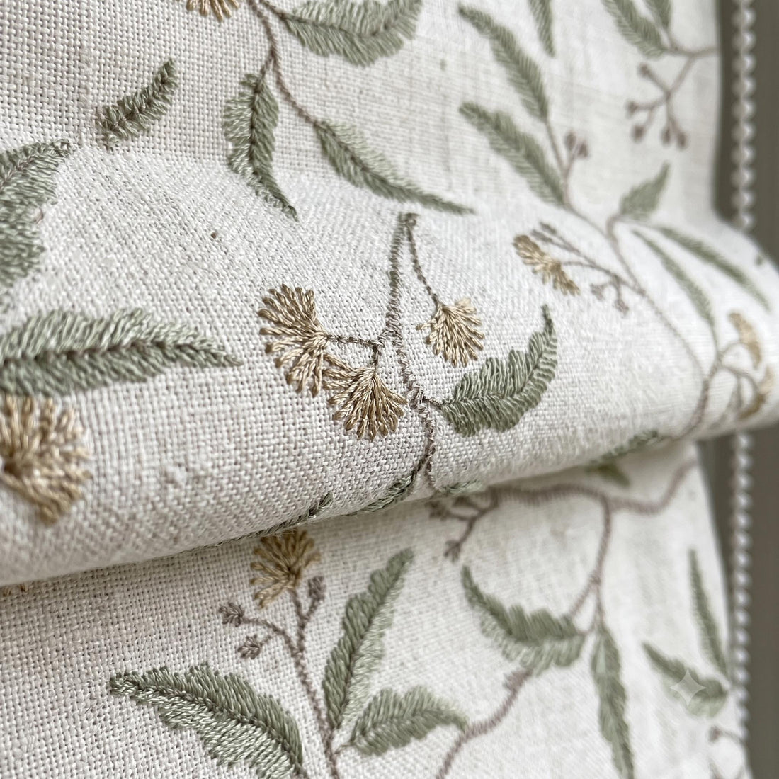 Close-up of ivory linen Roman shade folds with sage green leaves, taupe vines, and beige floral embroidery.